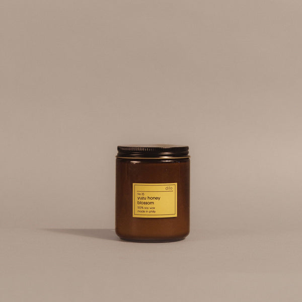 An amber glass jar candle with a black metal lid and a yellow label reading 'dis. beeswax honey blossom 100% beeswax' on a warm taupe background.
