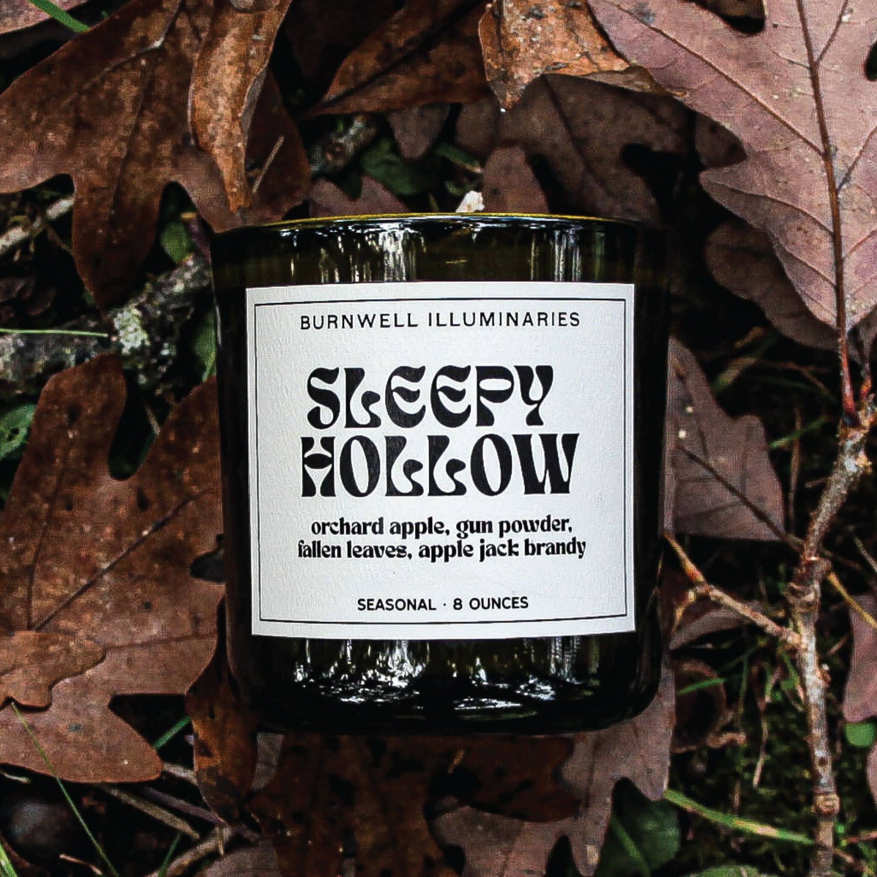Candle labeled 'Sleepy Hollow' by Burnwell Illuminaries on a bed of leaves.