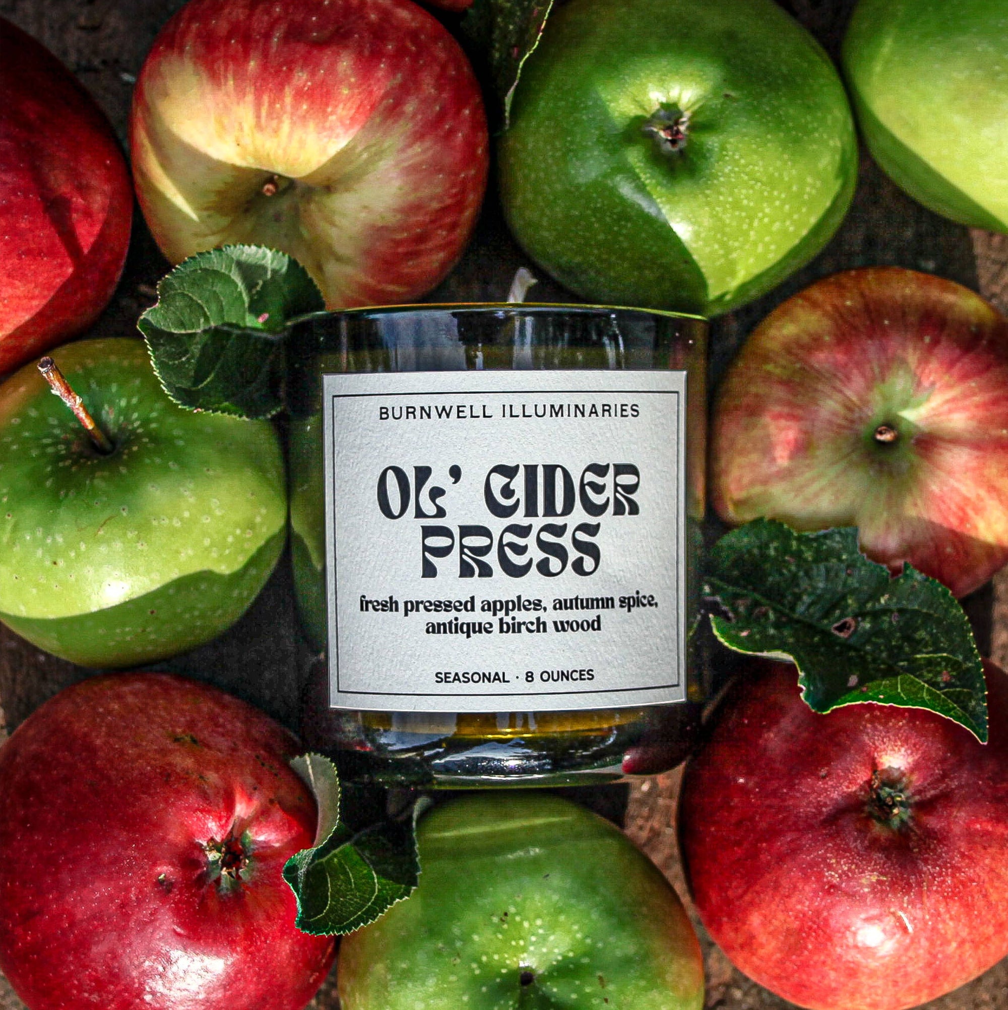 Candle labeled 'Old Cider Press' surrounded by red and green apples on a wooden surface.