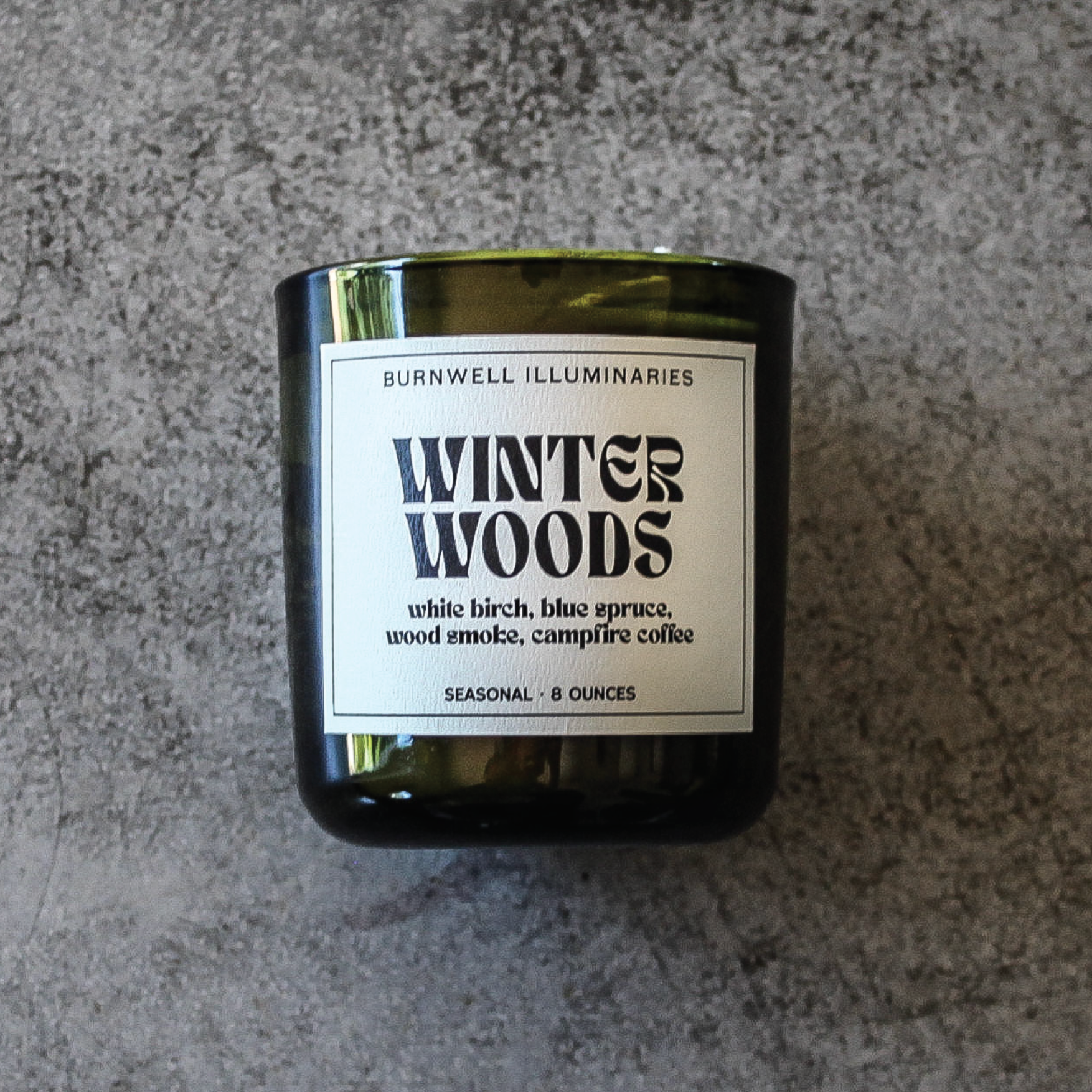 Candle labeled 'Winter Woods' by Burnwell Illuminaries on a gray surface