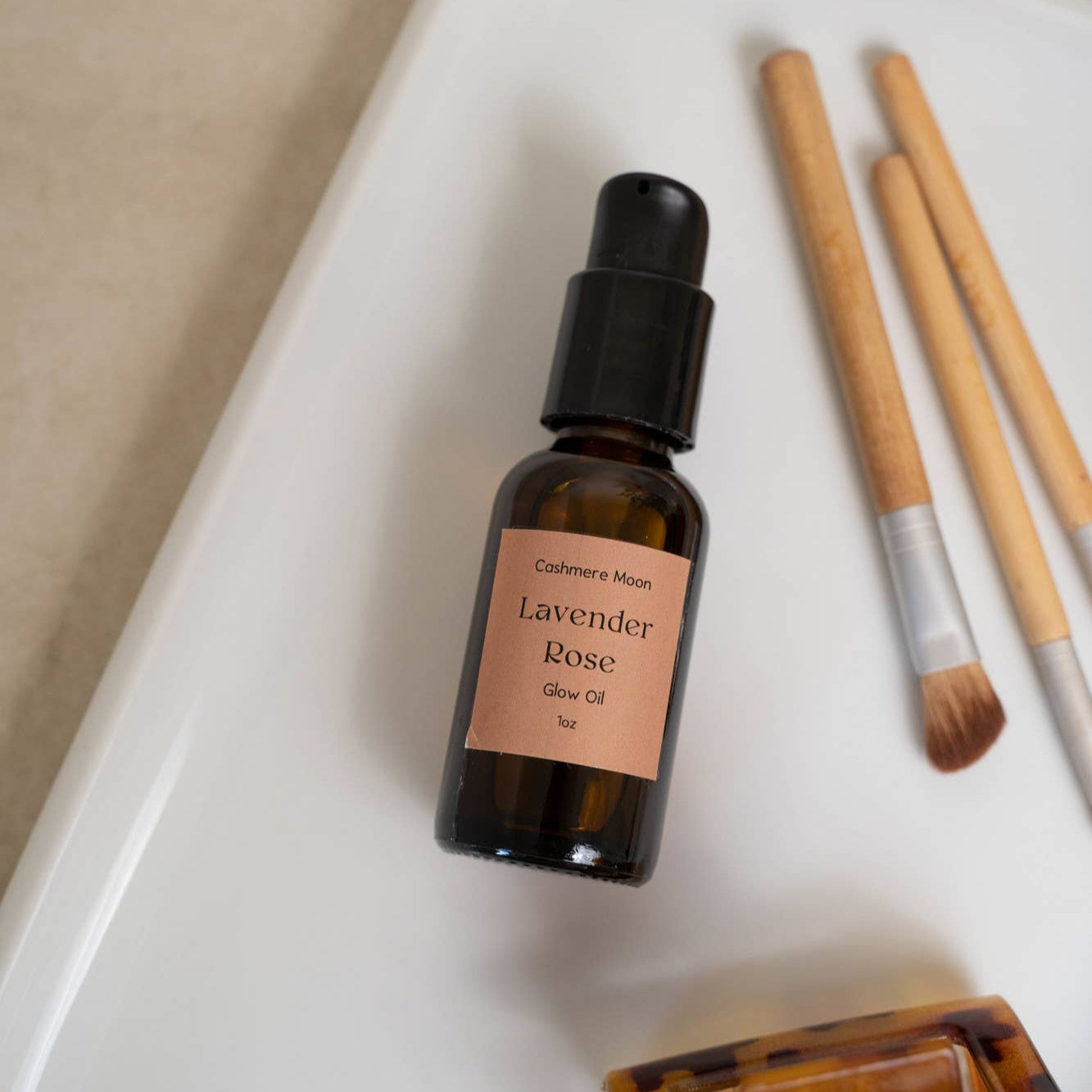 A travel-sized bottle of Lavender Rose Glow Oil with a dropper, placed on a white surface next to some bamboo brushes and a hair clip.