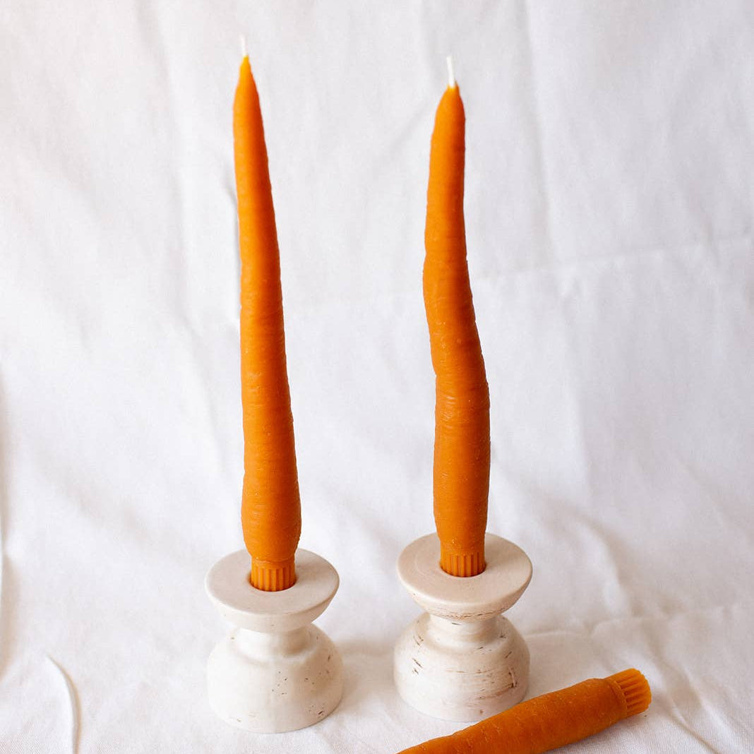 Two tall carrot candles in white candle holders with a carrot on a white background