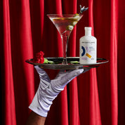 Hand in a white glove holding a tray with a cocktail and a bottle of Brightland oil against a red curtain background.