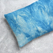 A rectangular eye pillow with a blue and white tie-dye pattern, resting on a textured gray concrete surface.
