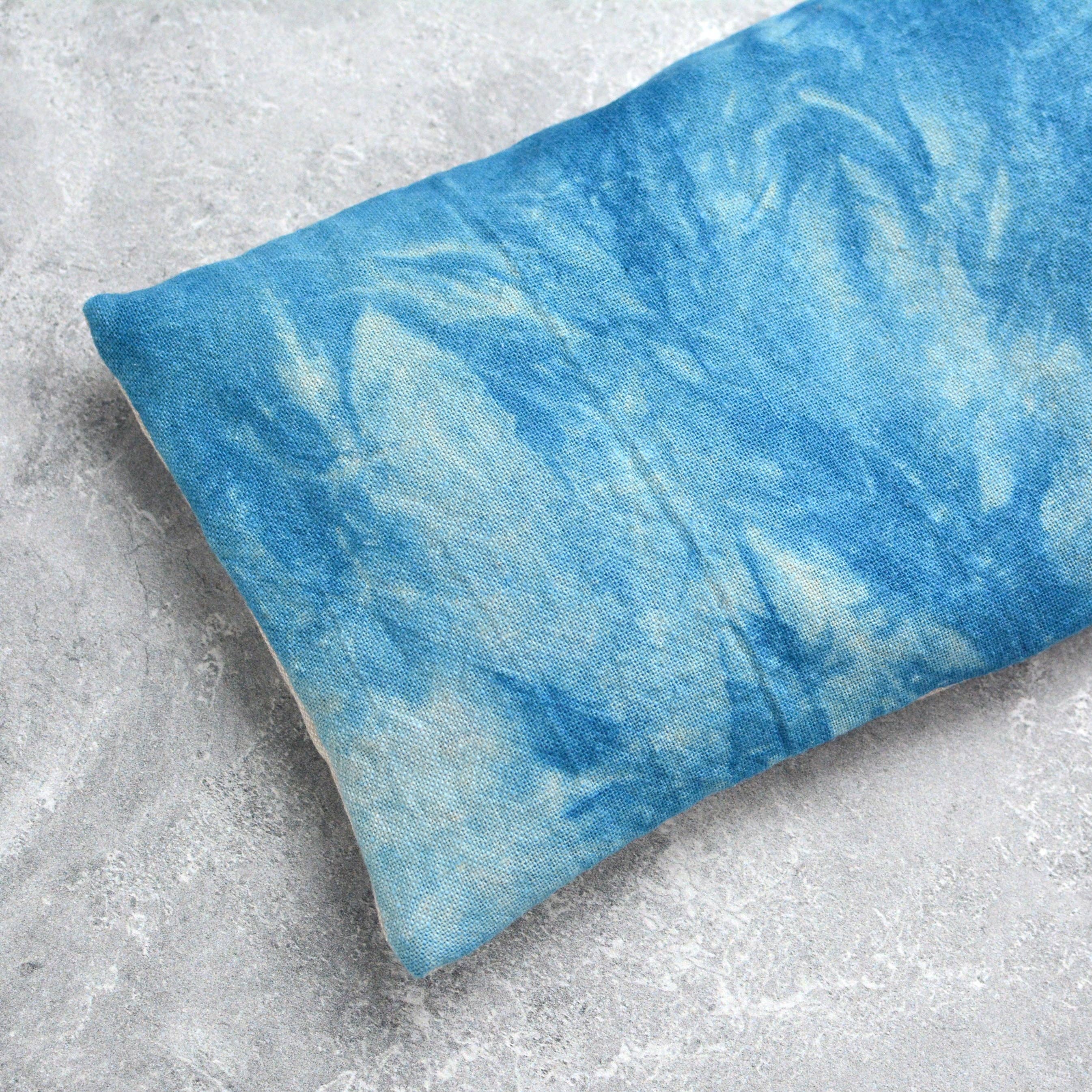 A rectangular eye pillow with a blue and white tie-dye pattern, resting on a textured gray concrete surface.