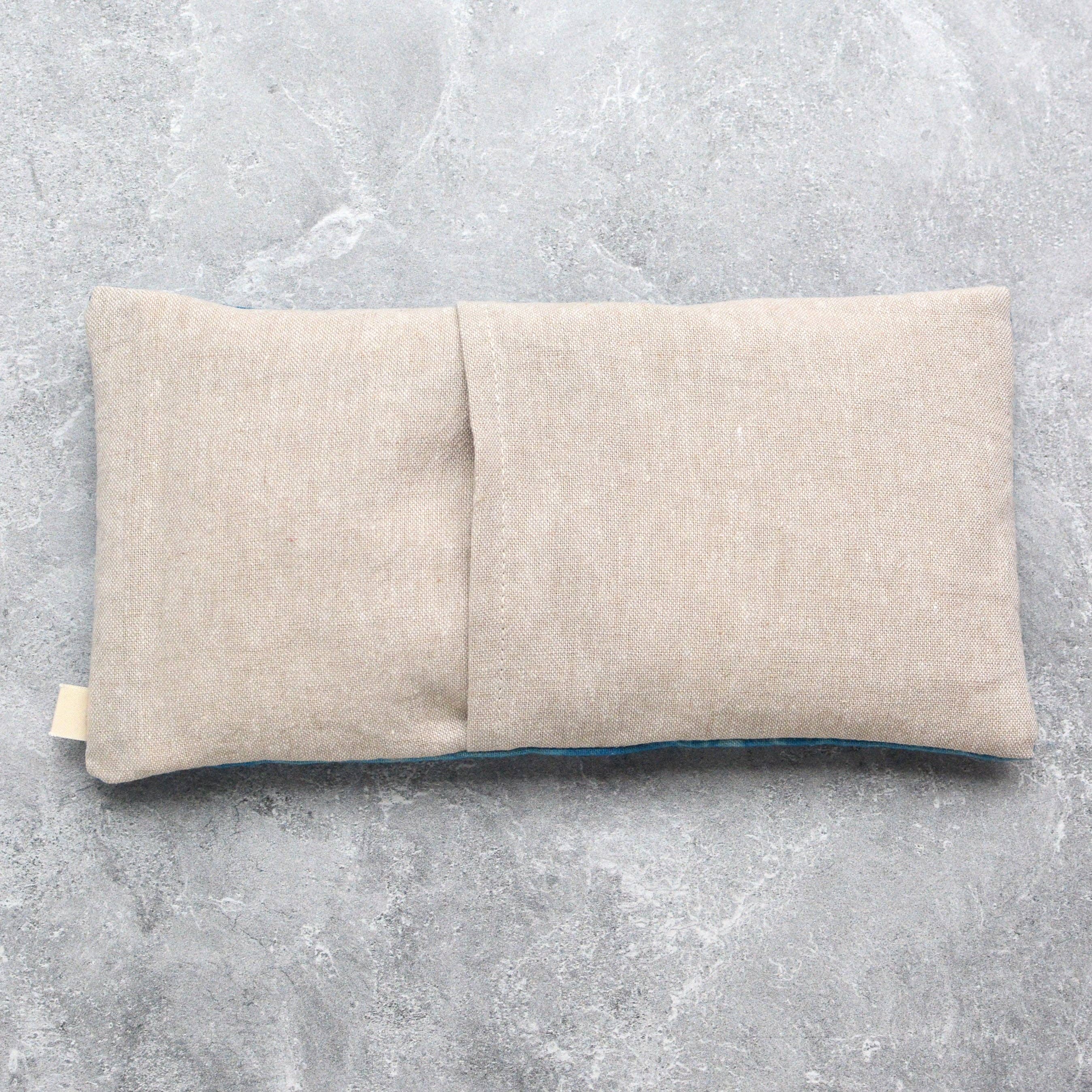 A rectangular beige linen eye mask pillow with a central fold, shown on a textured gray concrete surface.