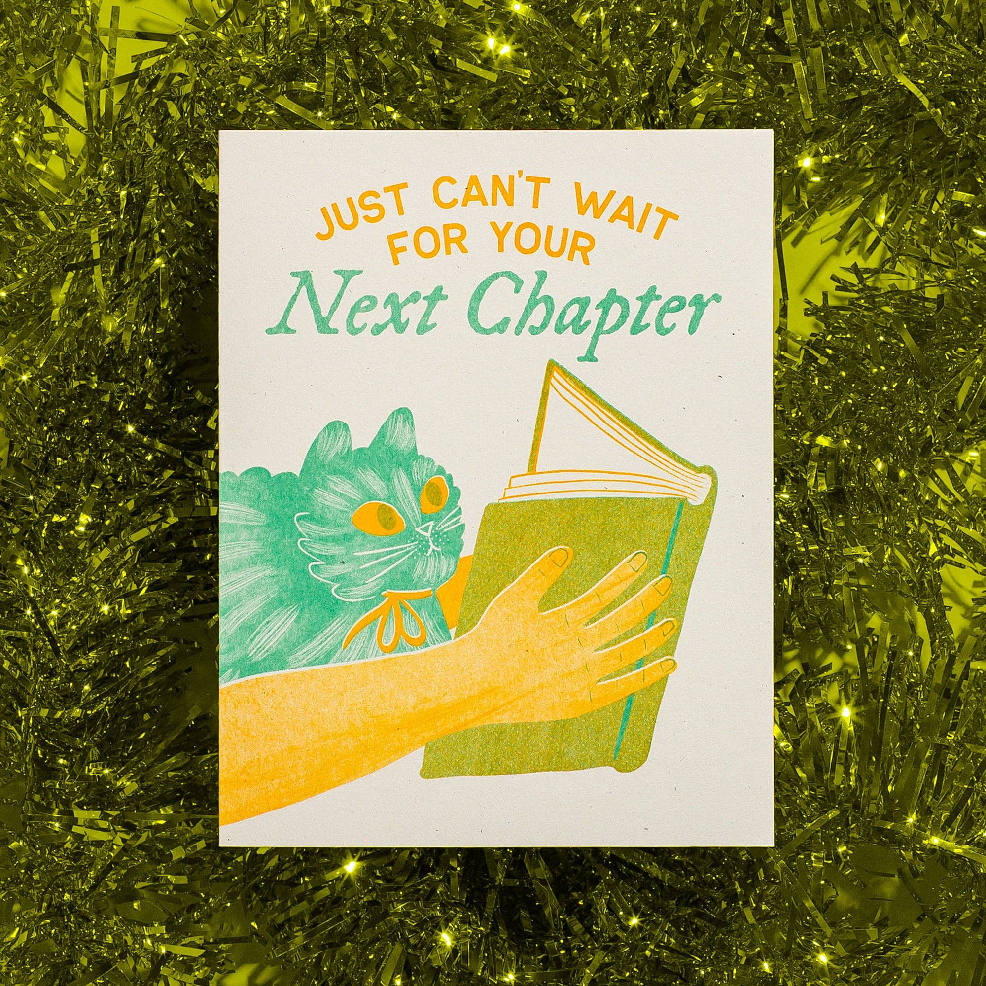 Card with a cat and hand holding a book, text 'Just can't wait for your next chapter' on a green textured background.