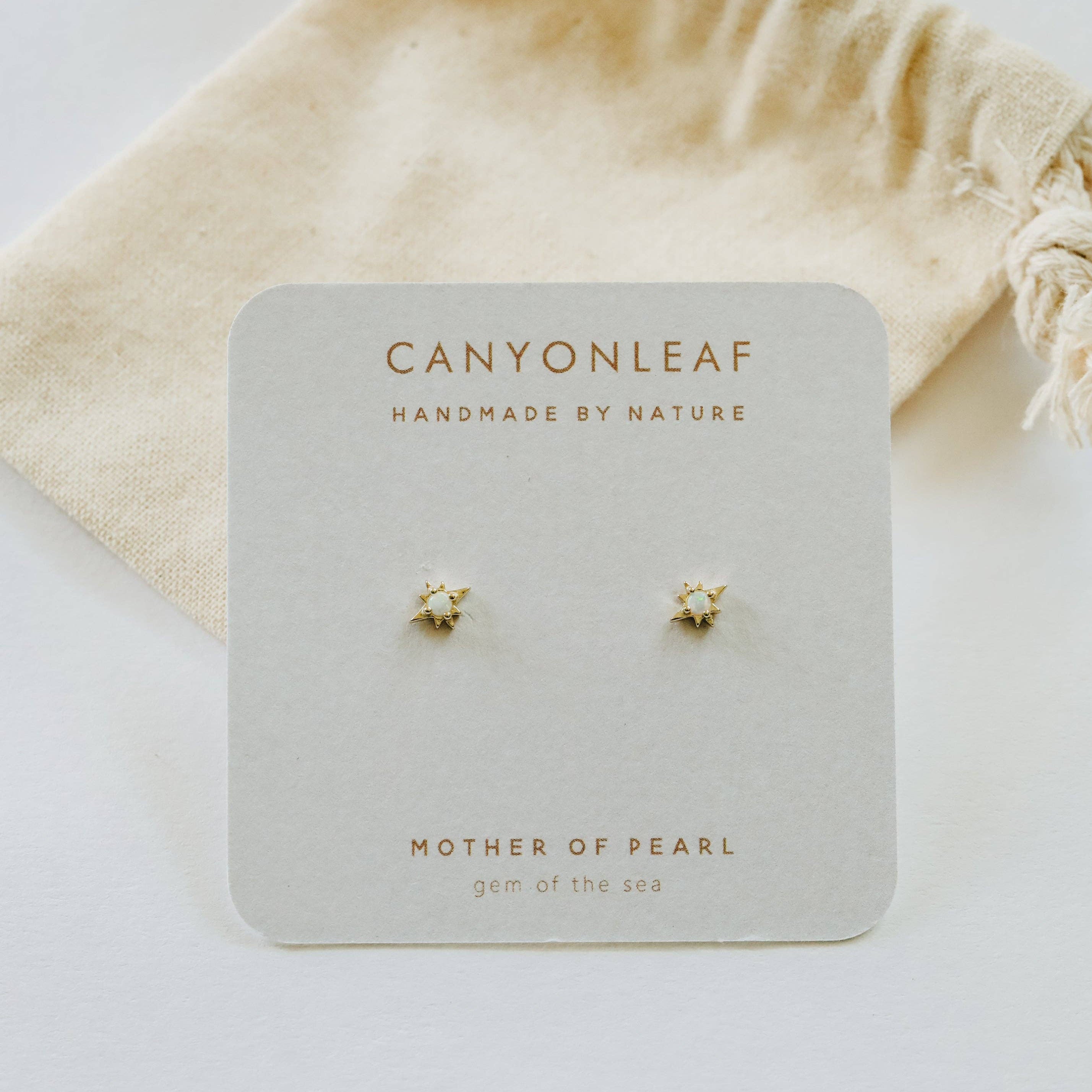 Pair of gold earrings on a Canyonleaf branded card with a beige drawstring bag in the background.