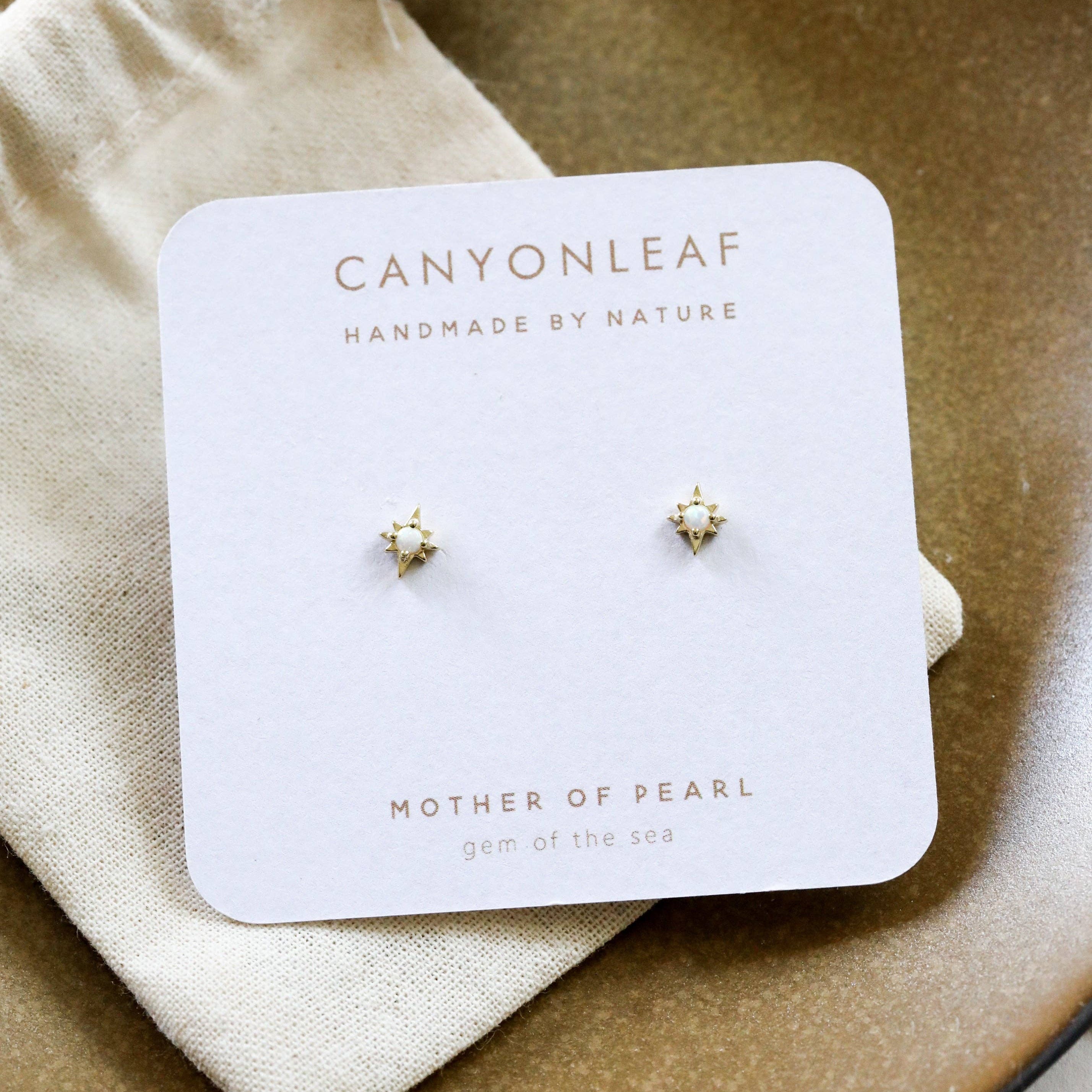 Pair of earrings on a Canyonleaf branded card with 'Mother of Pearl' text, displayed on a beige fabric and ceramic plate.