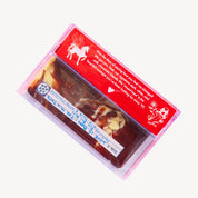 A pink translucent cassette-style case containing a brown chocolate bar, with a bright red printed label featuring white text and illustrations, photographed on a plain light background.