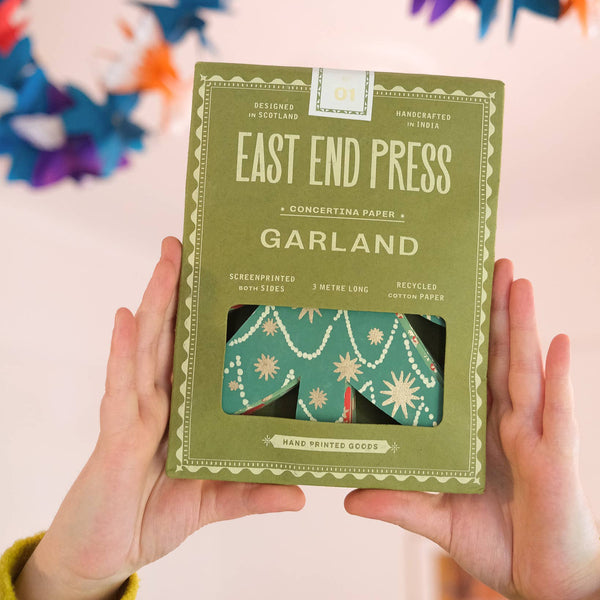 Green packaging of East End Press Garland held by hands with colorful flowers in the background