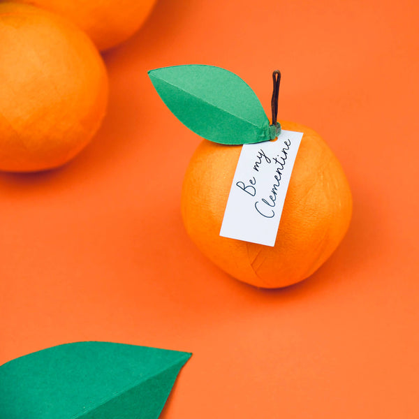 Decorative orange surprise ball with a green leaf and 'Be my Clementine' tag on an orange background