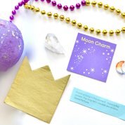 A flat-lay of a moon charm craft kit on a white background, featuring a gold foil crown, a purple card reading 'Moon Charm' with gold stars, a blue fortune card, a purple glitter ball, a clear crystal, and gold and purple bead strands.