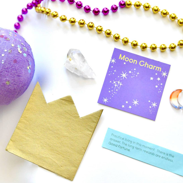 A flat-lay of a moon charm craft kit on a white background, featuring a gold foil crown, a purple card reading 'Moon Charm' with gold stars, a blue fortune card, a purple glitter ball, a clear crystal, and gold and purple bead strands.