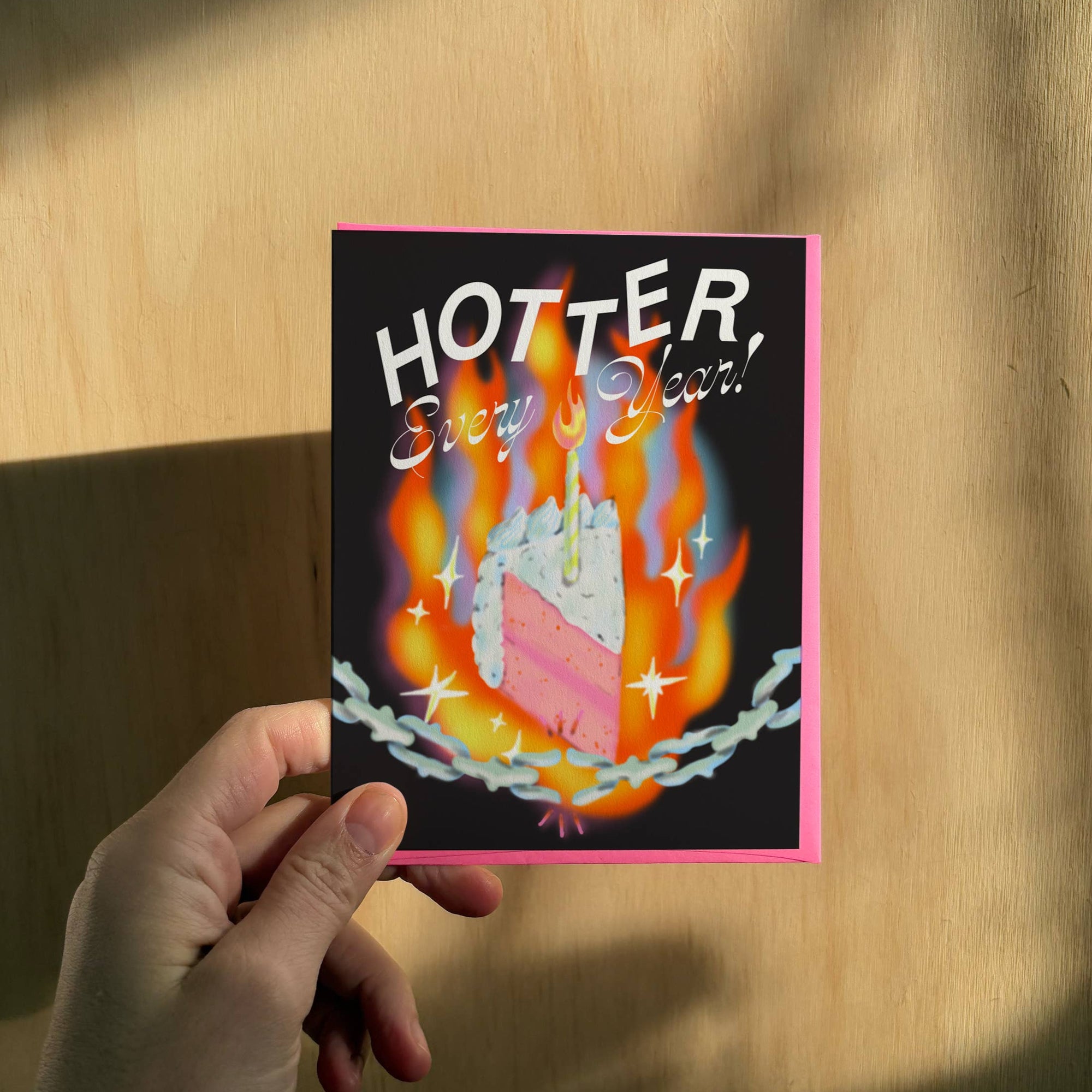 Hand holding a card with a birthday cake design and 'HOTTER Every Year!' text against a wooden background.