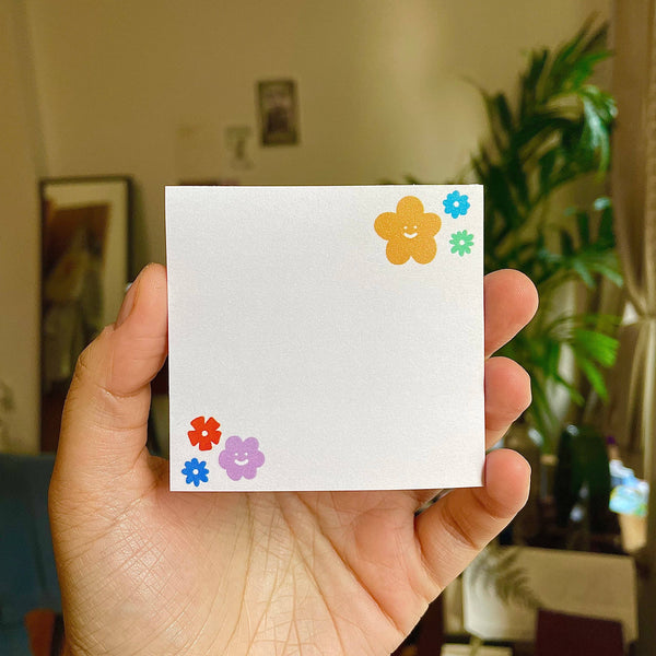 Hand holding a small white notepad with colorful flower stickers in an indoor setting.