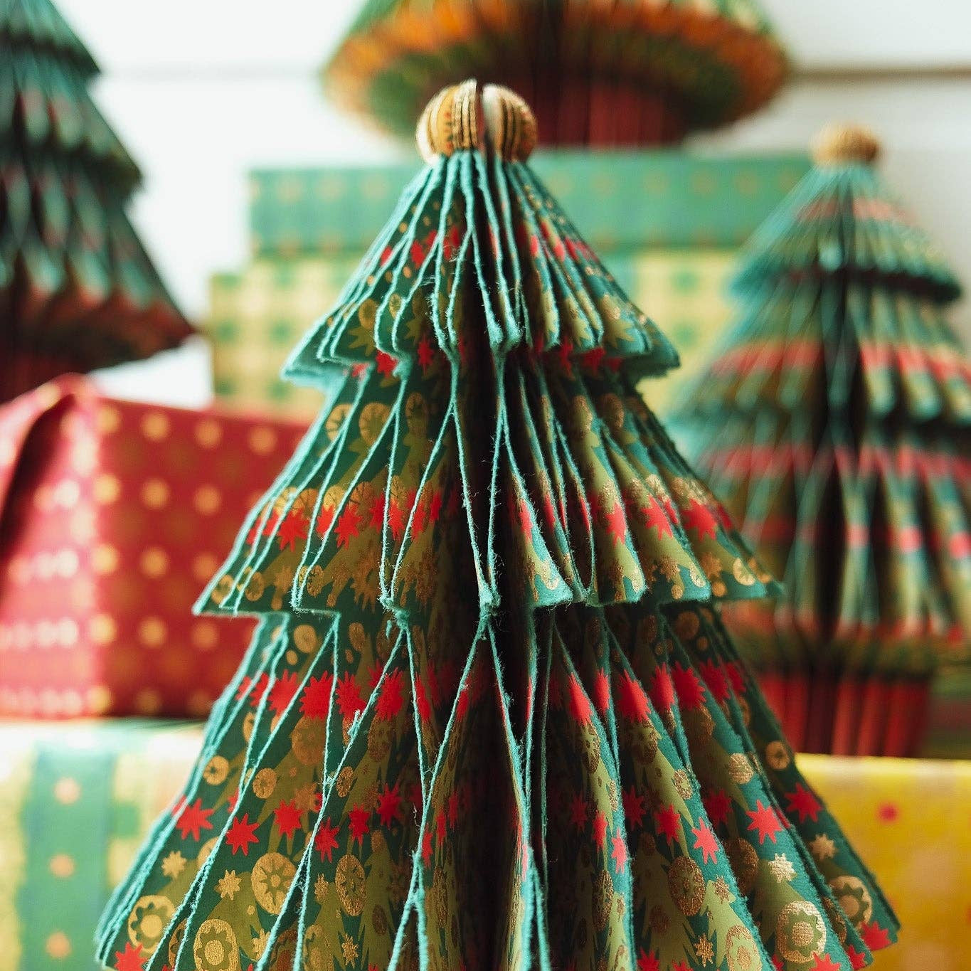 A decorative Christmas tree made from layered, pleated paper with a green, red, and gold holiday pattern, standing on a wooden surface with wrapped gifts in the background.