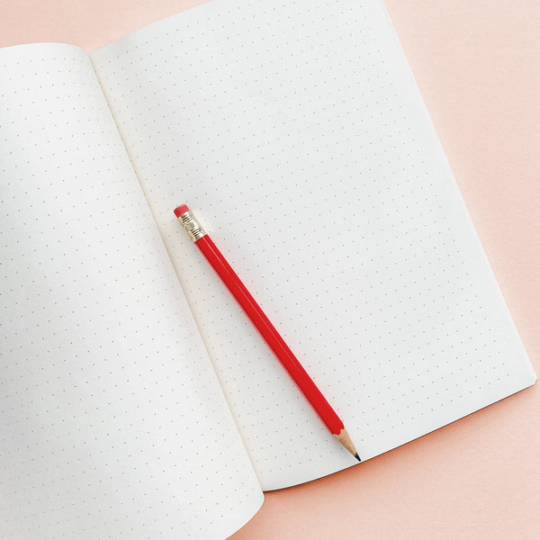 Red pencil resting on an open notebook with a dotted pattern against a pink background