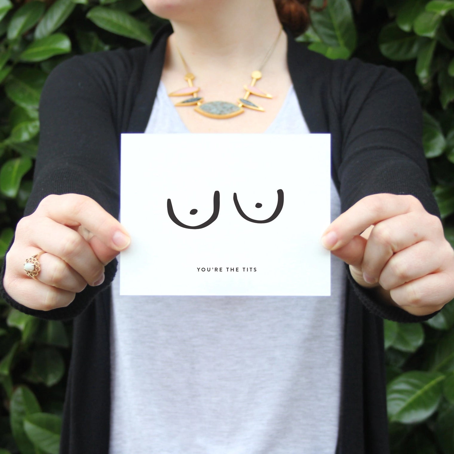 Person holding a card with illustrated breasts and and text that reads 'YOU'RE THE TITS' against a green leafy background