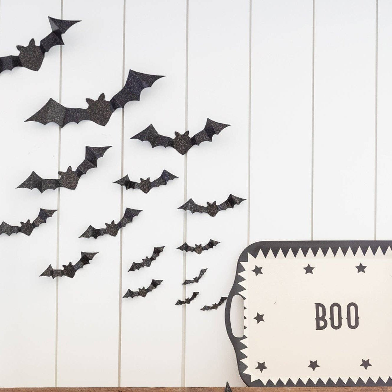Halloween wall decor featuring a flock of black bat cutouts on a white shiplap wall, with a white and black 'BOO' sign, two witch hats, and two small Christmas trees at the bottom right.