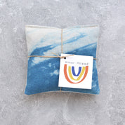 A square fabric lavender pillow wrapped in twine with a paper tag that reads 'minor thread' and shows a small rainbow logo, placed on a textured gray surface.