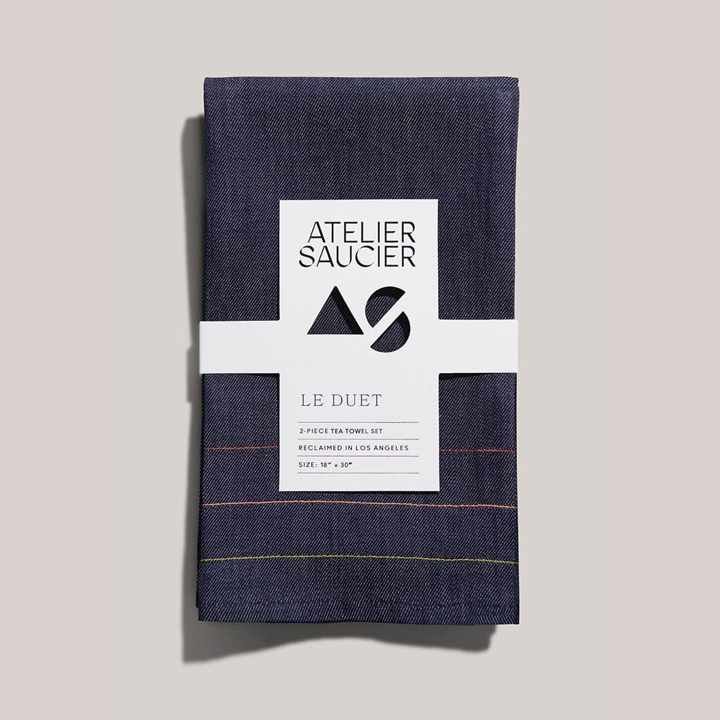 Two navy blue denim tea towels with a white label on a light gray background