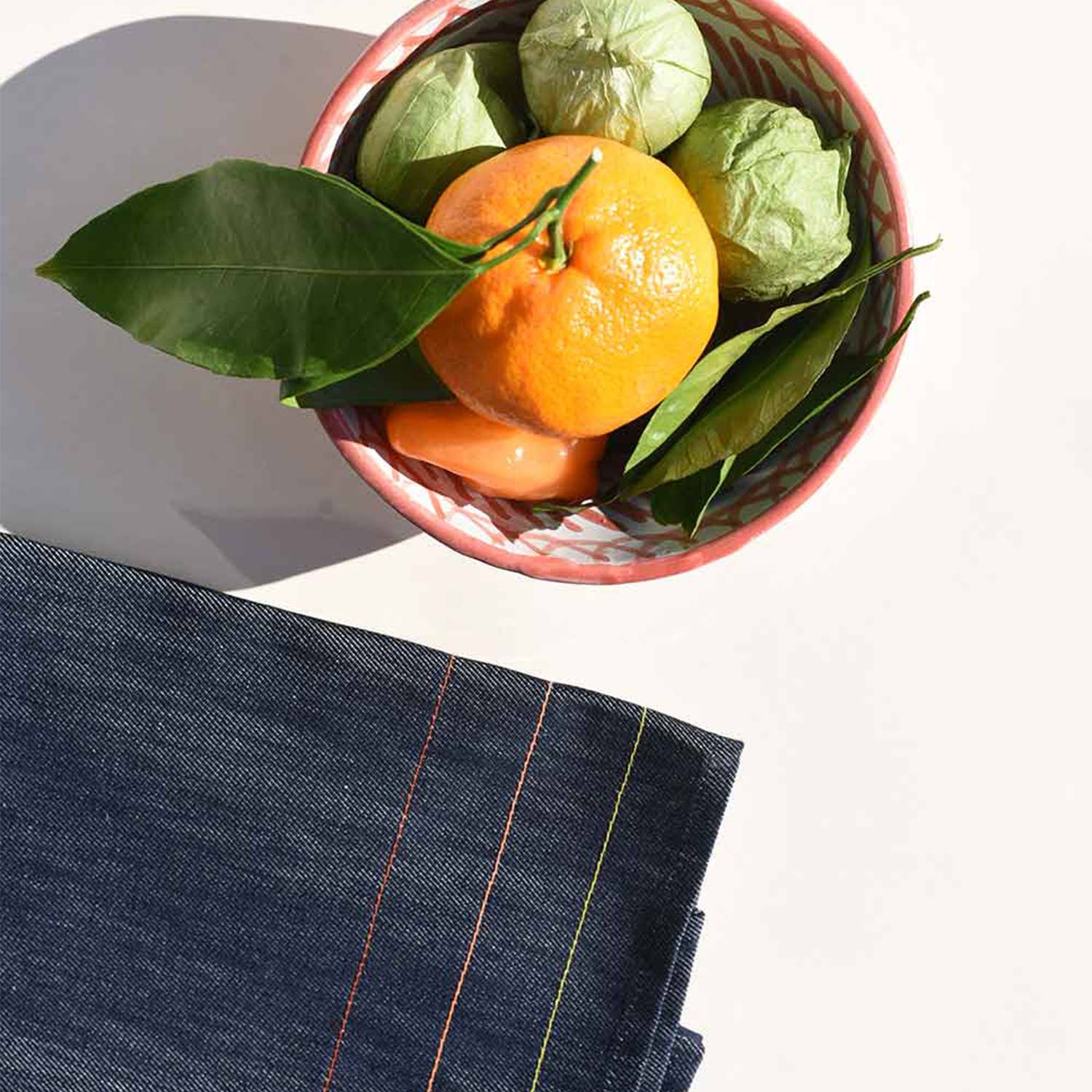 Bowl of oranges with green leaves on a white surface next to a denim fabric.