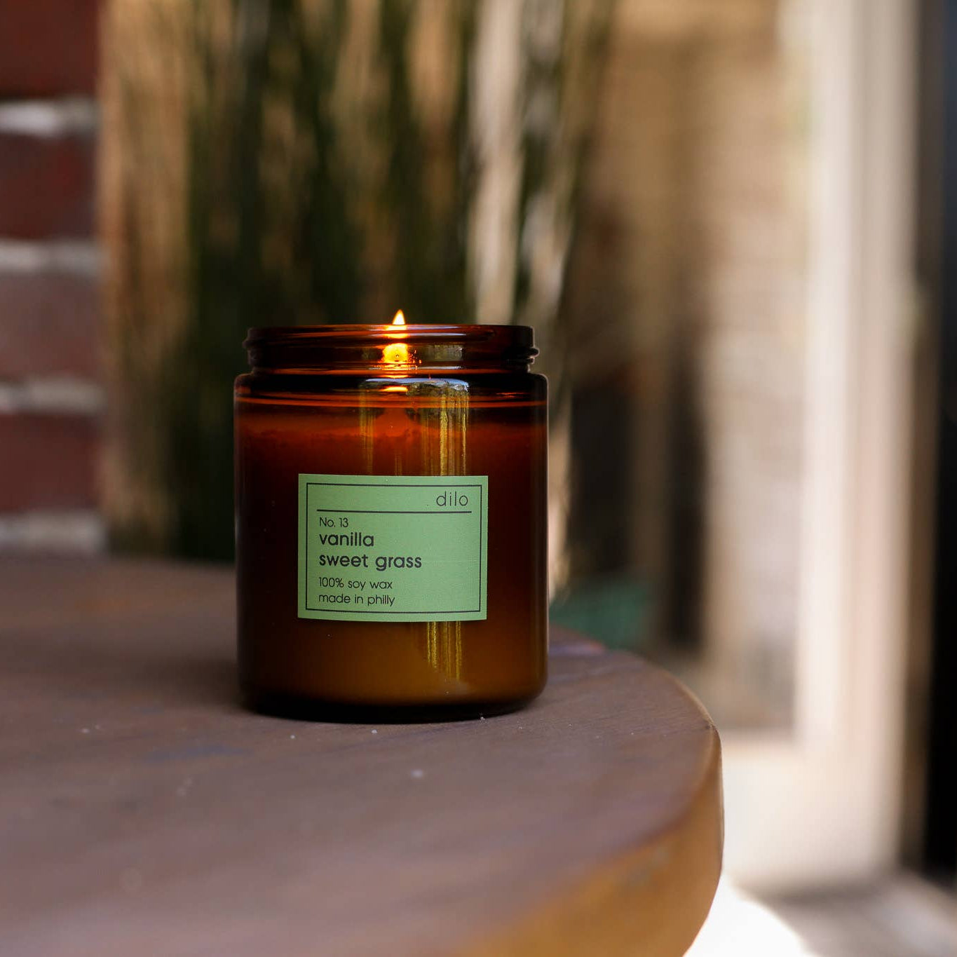 A vanilla sweet grass candle with a green label, placed on a wooden surface, with a brick wall and greenery in the background.