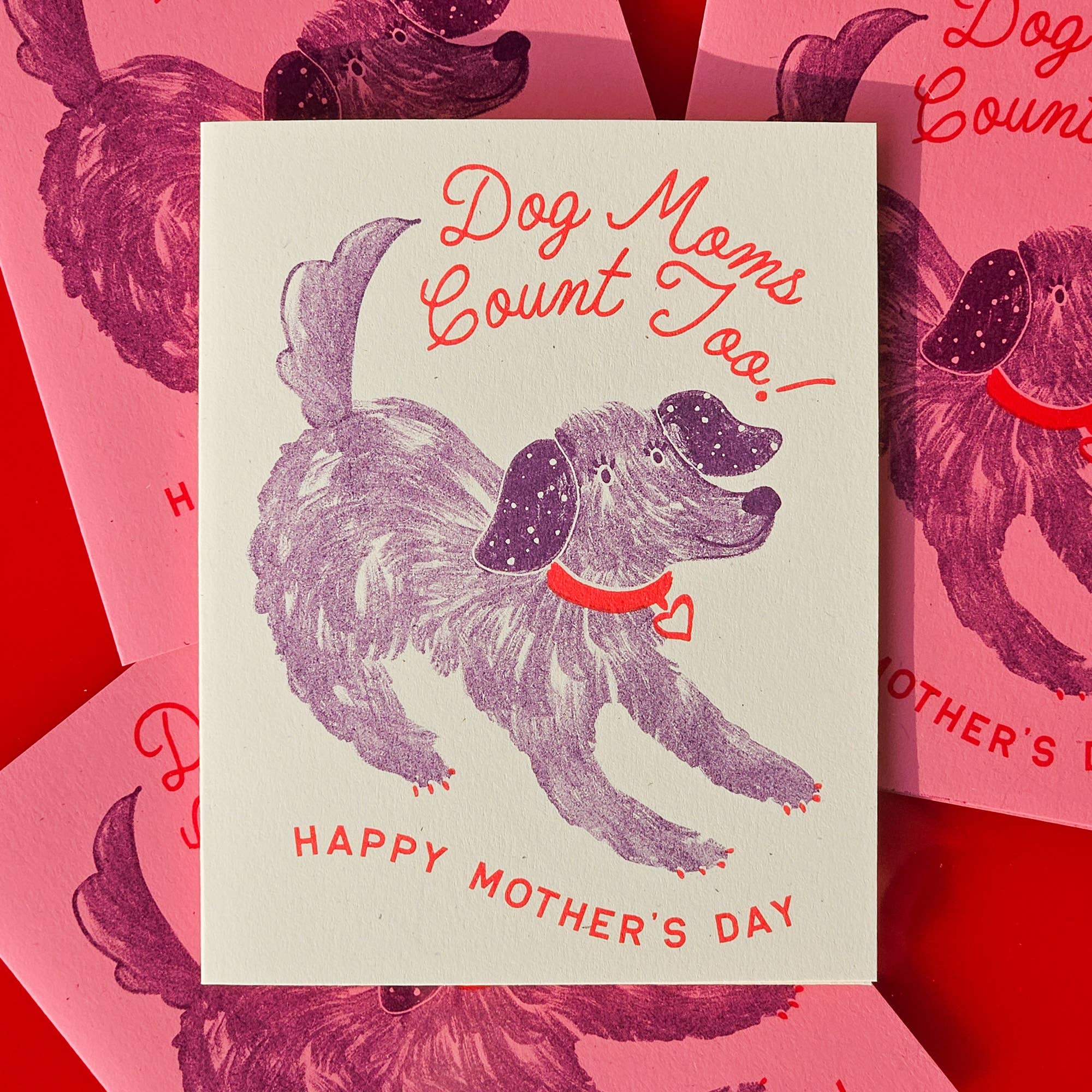A folded Mother's Day card with a risograph print of a dog and the text 'Dog Moms Count Too! Happy Mother's Day', displayed against a red background with similar images.