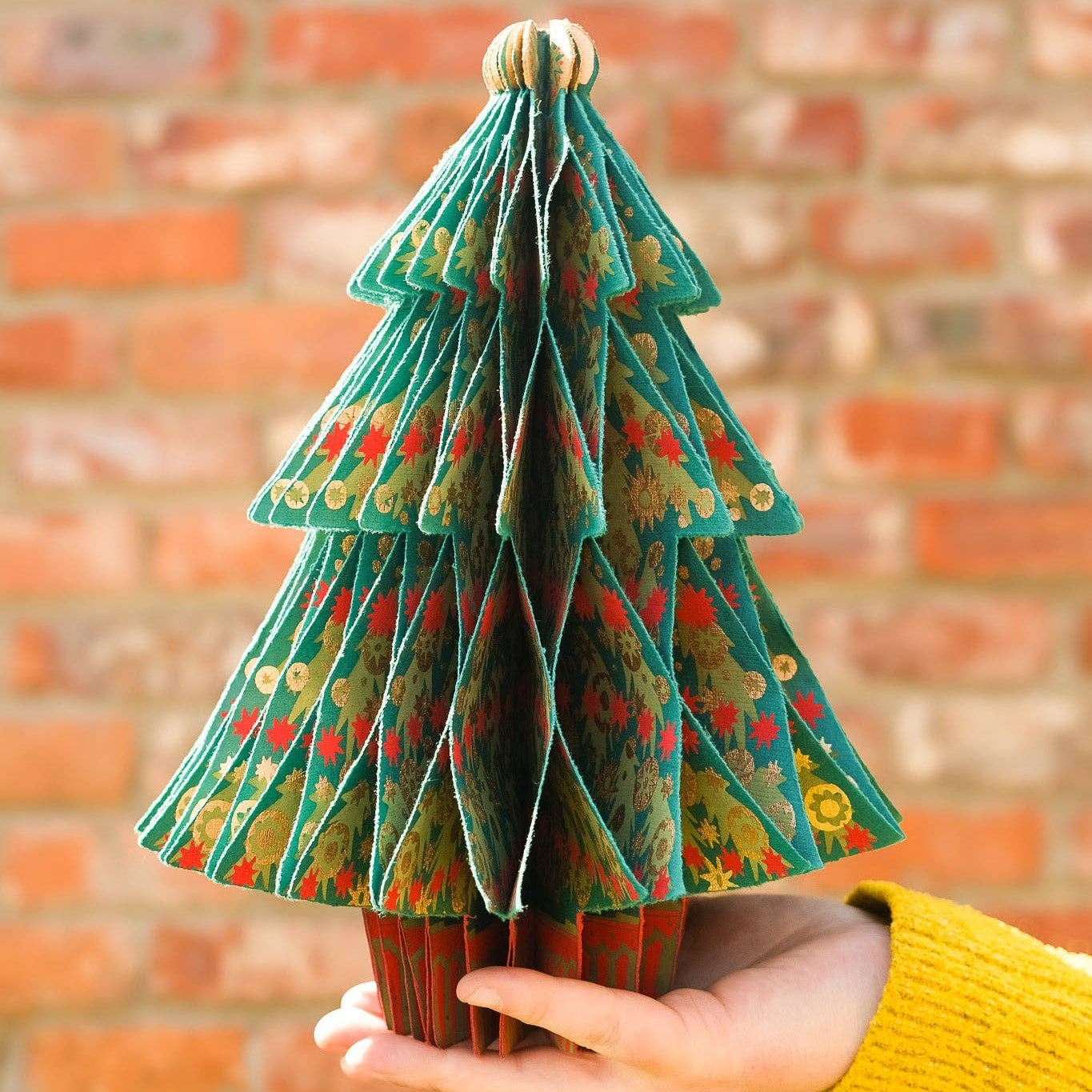 A small decorative Christmas tree made from layered green patterned paper, held in a person's hand against a blurred red brick wall background.