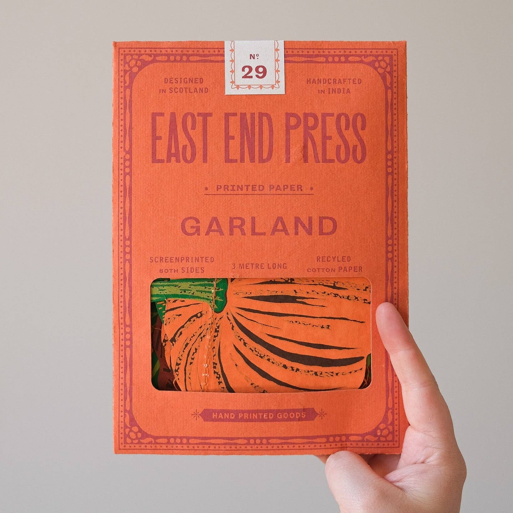 A hand holding a bright orange book titled 'EAST END PRESS GARLAND' against a plain light gray background. The cover features a window showing printed pumpkin artwork, and a small label at the top reads 'No.