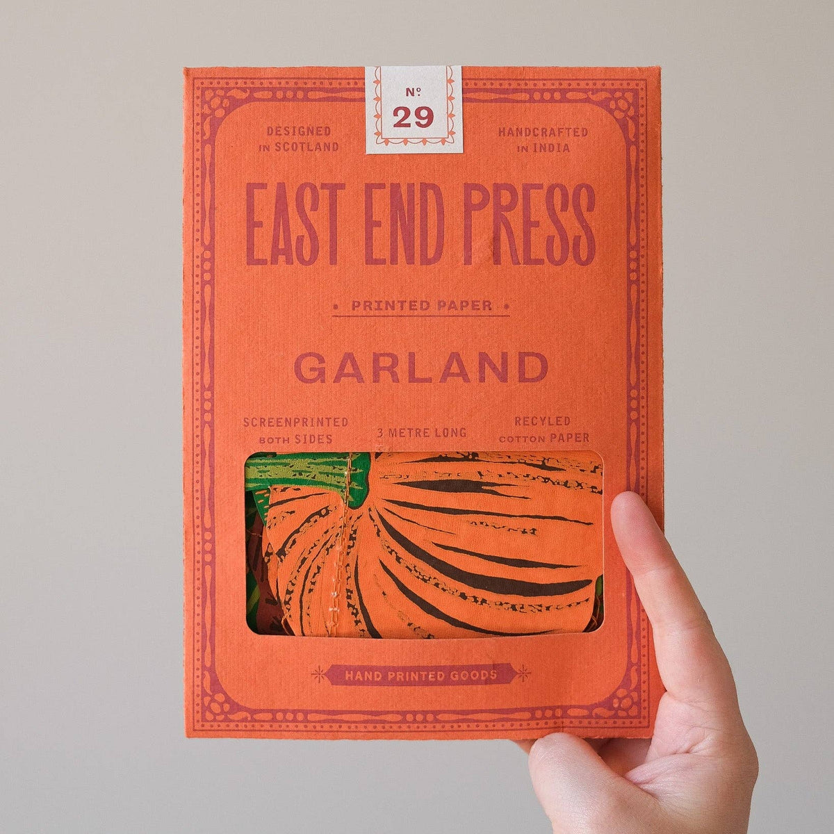 A hand holding a bright orange book titled 'EAST END PRESS GARLAND' against a plain light gray background. The cover features a window showing printed pumpkin artwork, and a small label at the top reads 'No.