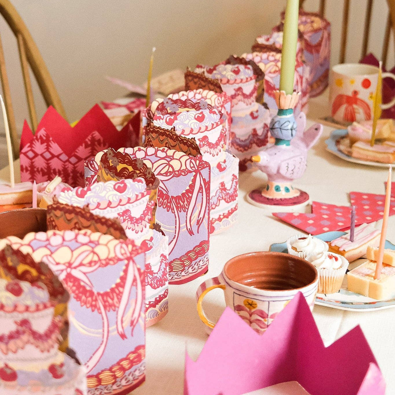 A styled tabletop display featuring multiple paper crown party hats in pink, red, and white patterns, along with matching cups, candles, and small cakes on a table.