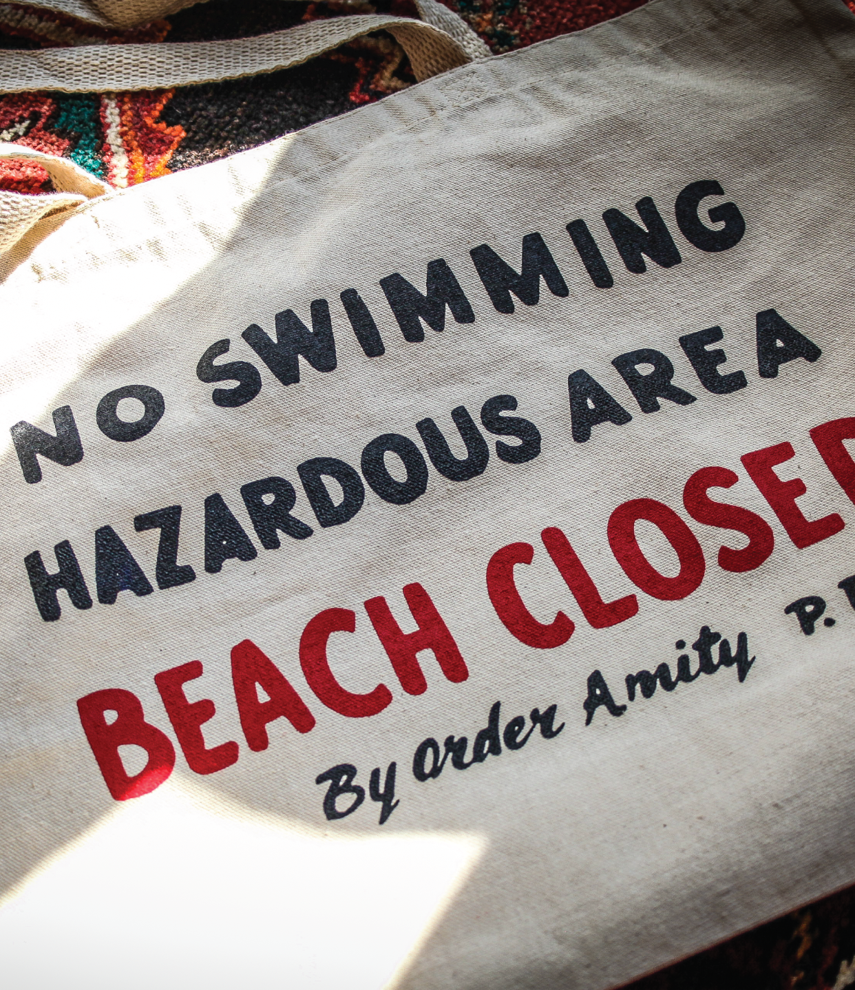Text sign reading 'No Swimming Hazardous Area Beach Closed By Order Amity P.' on a fabric background.