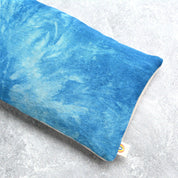 A rectangular blue textured eye pillow with a marbled tie-dye pattern in shades of blue, resting on a light gray concrete-textured surface. A small white brand tag is visible at the bottom right corner.
