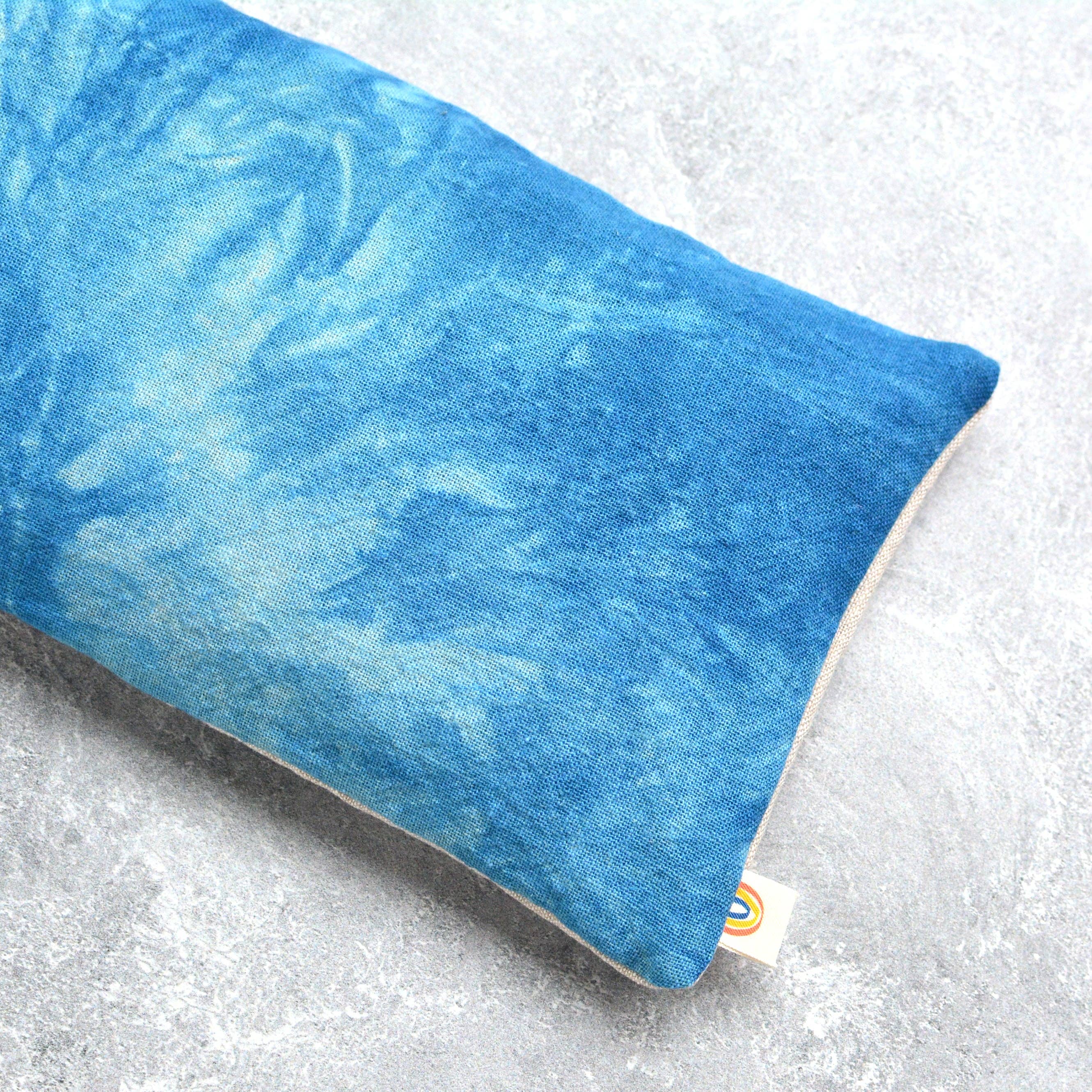 A rectangular blue textured eye pillow with a marbled tie-dye pattern in shades of blue, resting on a light gray concrete-textured surface. A small white brand tag is visible at the bottom right corner.
