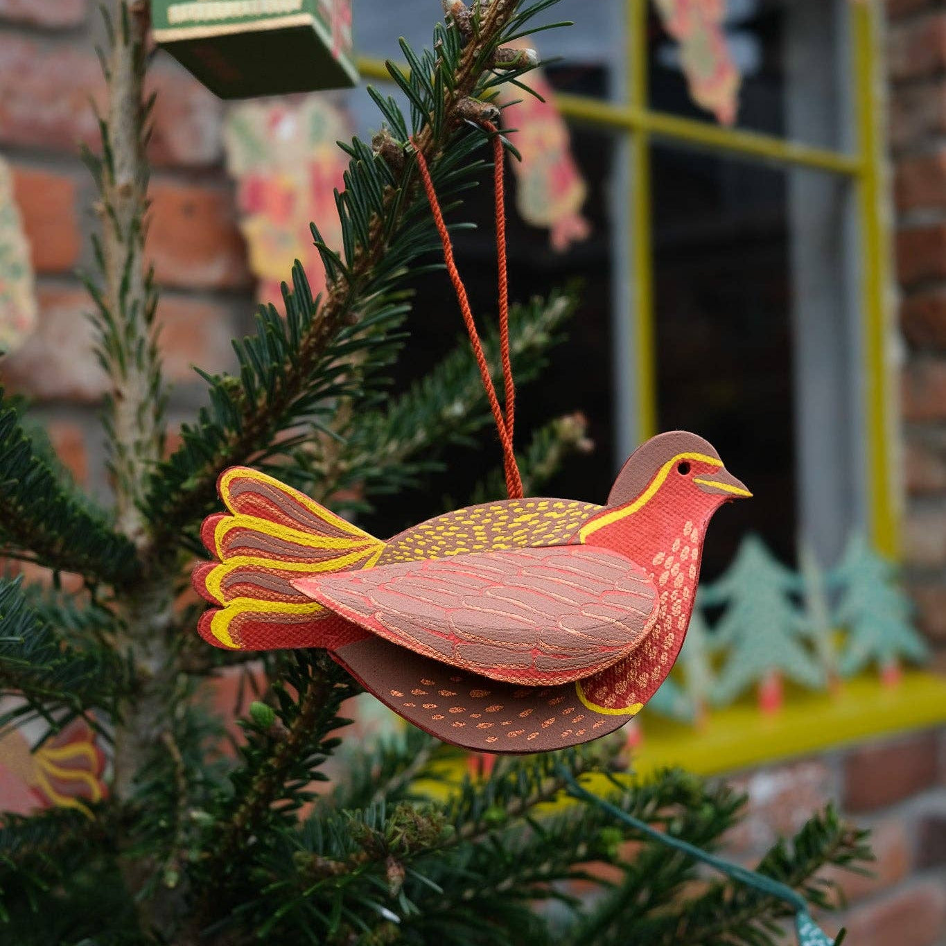 A screen printed paper ornament in the shape of a robin perched on a tree, with a house and window in the background, hung by a cotton loop.