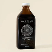 A dark amber glass bottle of Fat of the Land Elderberry Syrup with a white cap and black label, set against a solid light beige background.