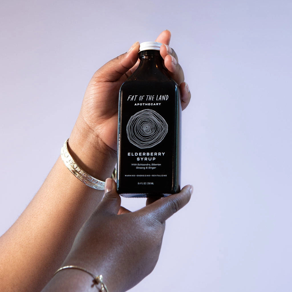 A person's hands holding a dark amber glass bottle of Fat of the Land Anniversary Elderberry Syrup against a solid light lavender background.
