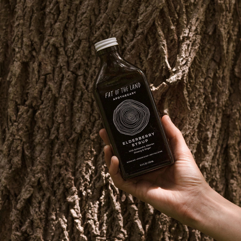 A hand holds a bottle of Fat of the Land Apothecary Elderberry Syrup in front of a textured tree trunk background.