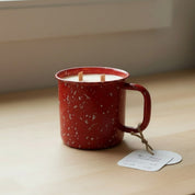 Red speckled mug with a candle inside on a wooden surface near a window.