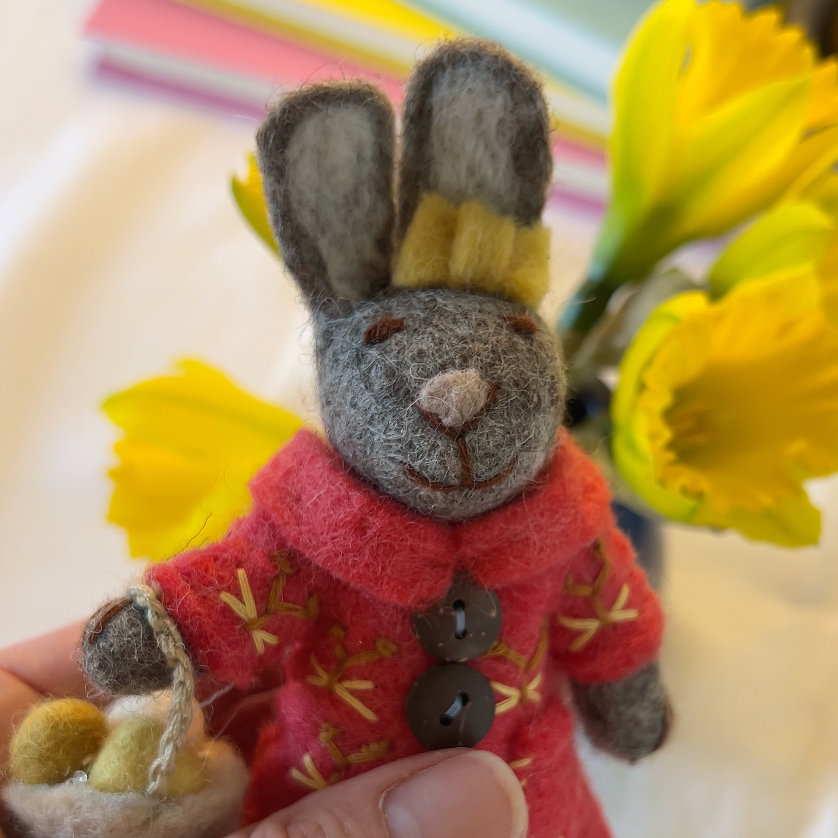 Small rabbit plush toy in a red coat held by a hand with flowers in the background