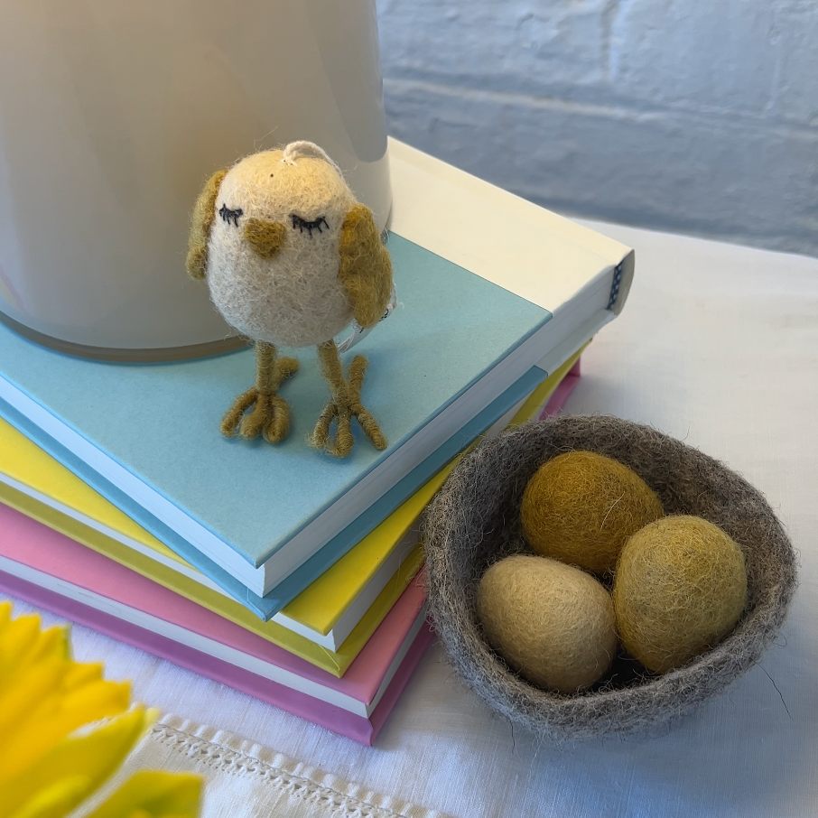 Small felt chicken on colorful books with a nest of felt eggs on a white surface.