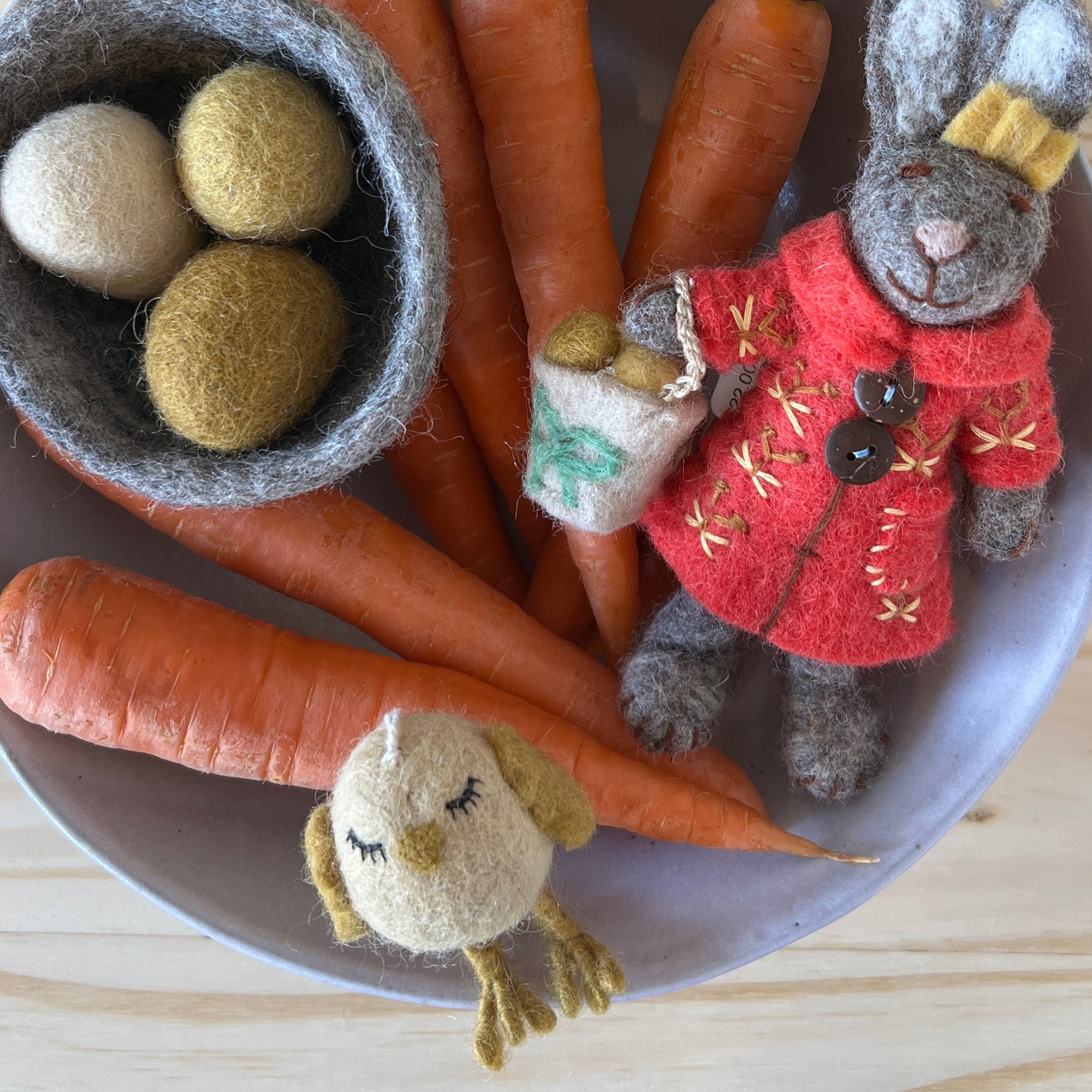 Decorative arrangement with carrots, a small rabbit figure, and a felt chicken on a wooden surface.