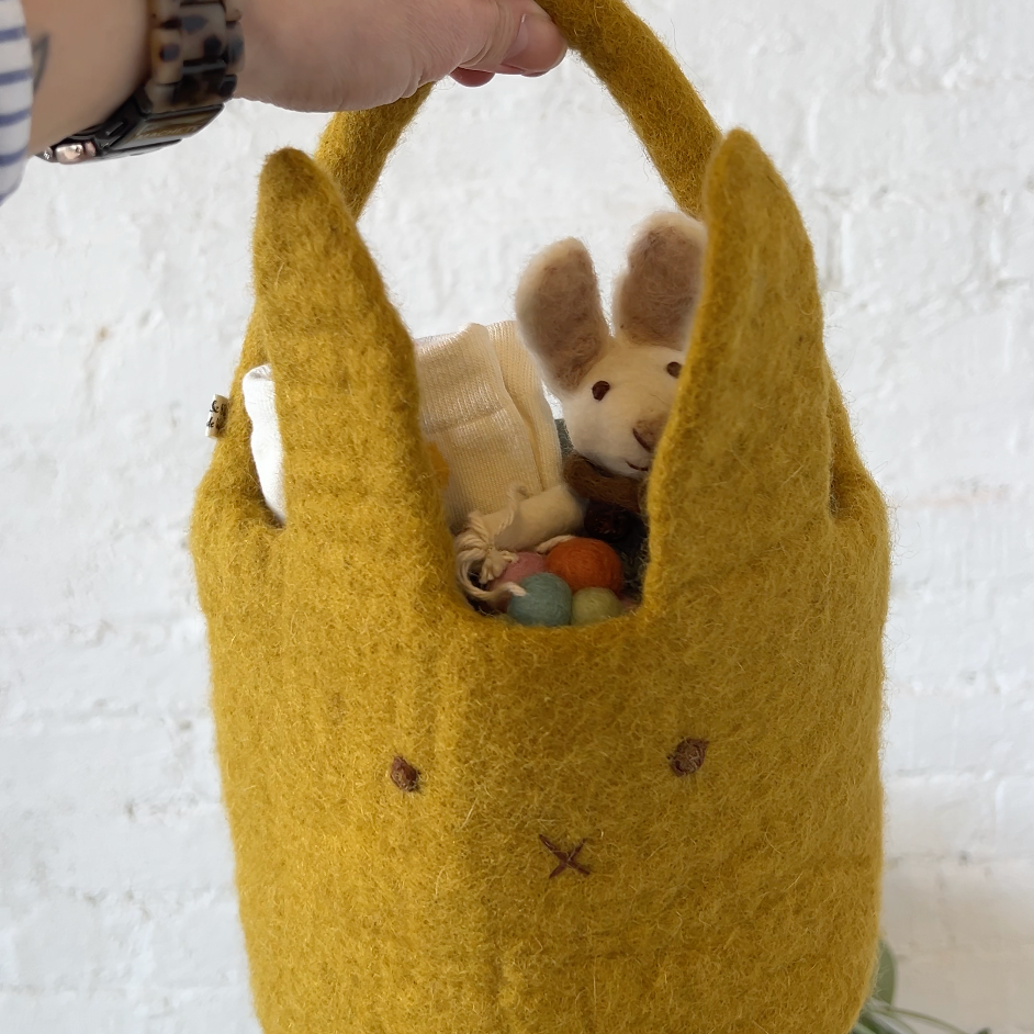 Yellow felted bag with a plush toy inside, held by a hand against a white textured wall.