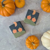 Two soap bars with pumpkin and moon design on a gray surface with pumpkins around