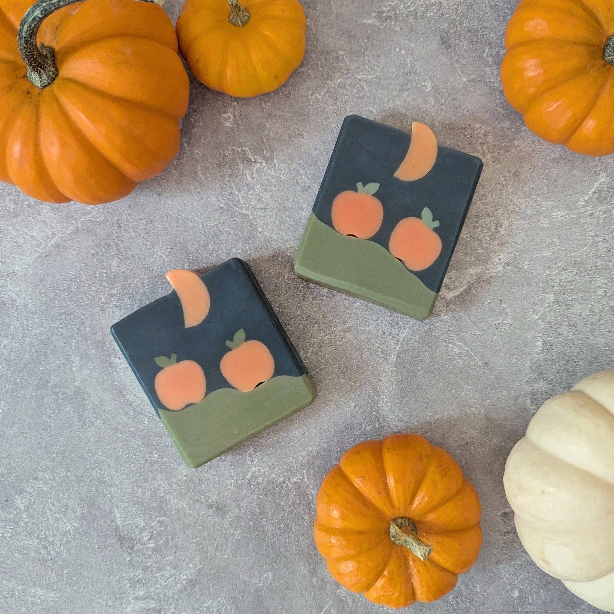 Two soap bars with pumpkin and moon design on a gray surface with pumpkins around