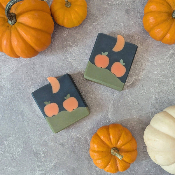 Two soap bars with pumpkin and moon design on a gray surface with pumpkins around