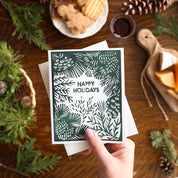 A hand holds a square holiday greeting card with a dark green and white pine-themed design and the words 'HAPPY HOLIDAYS' in the center, surrounded by a rustic holiday tabletop scene with pine branches, cookies, cheese, and a glass of red wine.