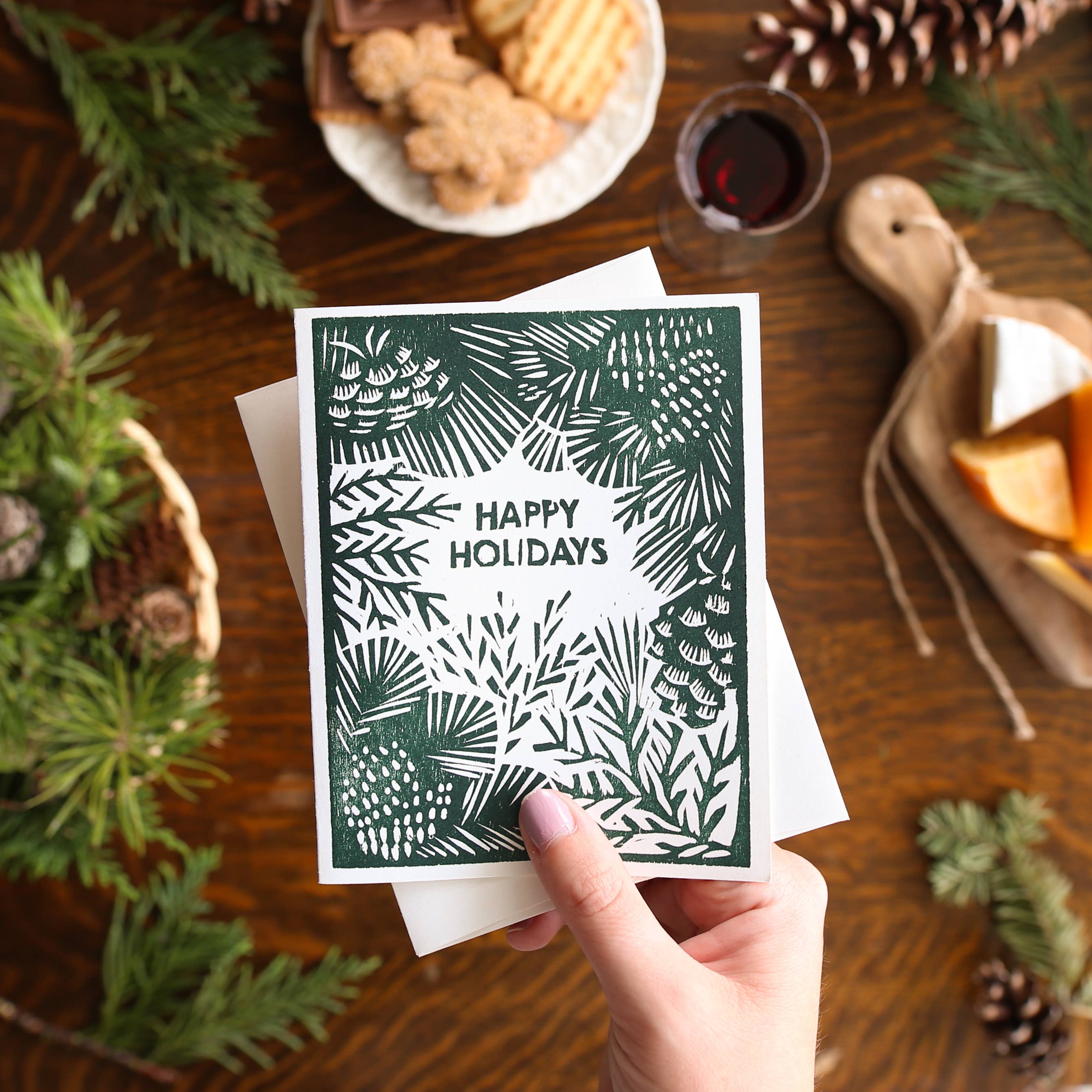 A hand holds a square holiday greeting card with a dark green and white pine-themed design and the words 'HAPPY HOLIDAYS' in the center, surrounded by a rustic holiday tabletop scene with pine branches, cookies, cheese, and a glass of red wine.